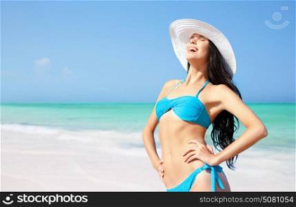 summer holidays, vacation, travel and people concept - smiling young woman in bikini swimsuit and sun hat on beach over sea and blue sky background. smiling young woman in sun hat on summer beach