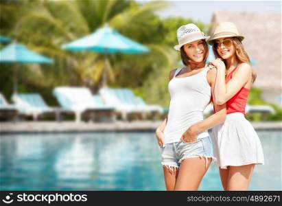 summer holidays, vacation, travel and people concept - smiling young women in hats and casual clothes over exotic beach with palm trees and pool background