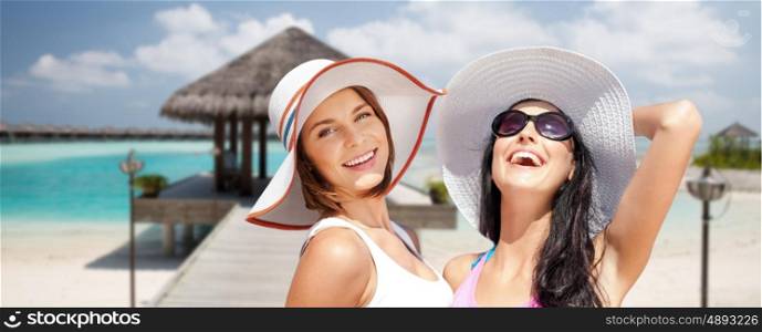 summer holidays, vacation, travel and people concept - smiling young women in hats and casual clothes over exotic tropical beach with bungalow shed background