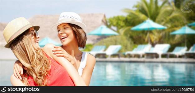 summer holidays, vacation, travel and people concept - smiling young women in hats and casual clothes over exotic beach with palm trees and pool background. smiling young women in hats on beach