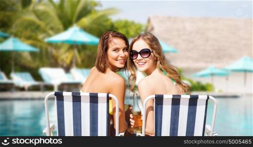 summer holidays, vacation, travel and tourism people concept - smiling young women with drinks sunbathing on chairs over swimming pool background. happy young women with drinks sunbathing on beach