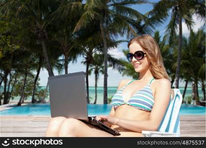 summer holidays, vacation, travel, technology and people concept - smiling women in sunglasses with laptop computer sunbathing in lounge over exotic tropical beach with palm trees and pool background
