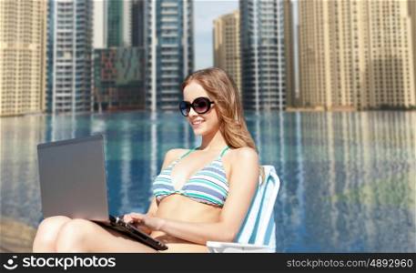 summer holidays, vacation, travel, technology and people concept - smiling women in sunglasses with laptop computer sunbathing in lounge over infinity edge pool in dubai background