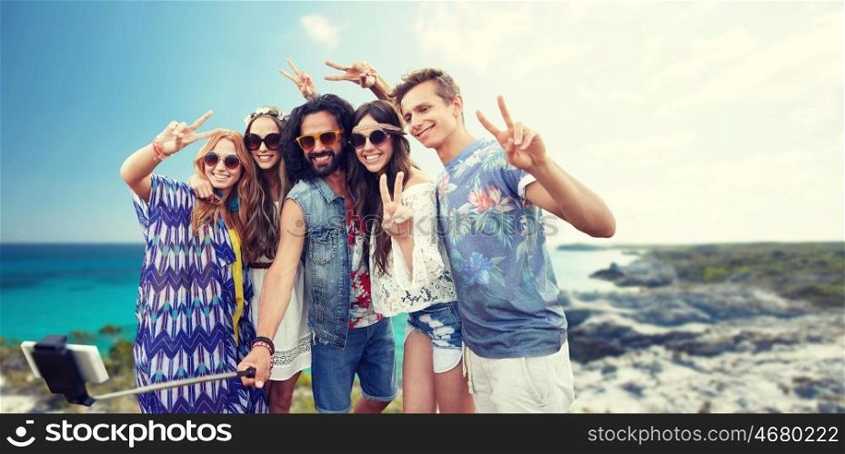 summer holidays, vacation, travel, technology and people concept - smiling young hippie friends taking picture by smartphone on selfie stick and showing peace gesture over beach background