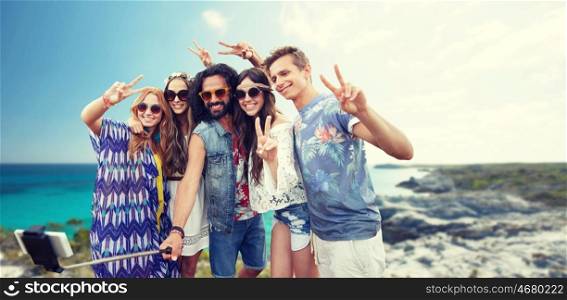 summer holidays, vacation, travel, technology and people concept - smiling young hippie friends taking picture by smartphone on selfie stick and showing peace gesture over beach background