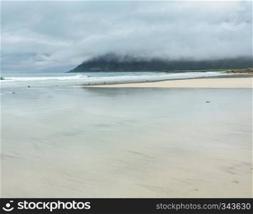 Summer misty and cloudy beach view with sky reflections in wave water covered white sand  Ramberg, Lofoten, Norway .