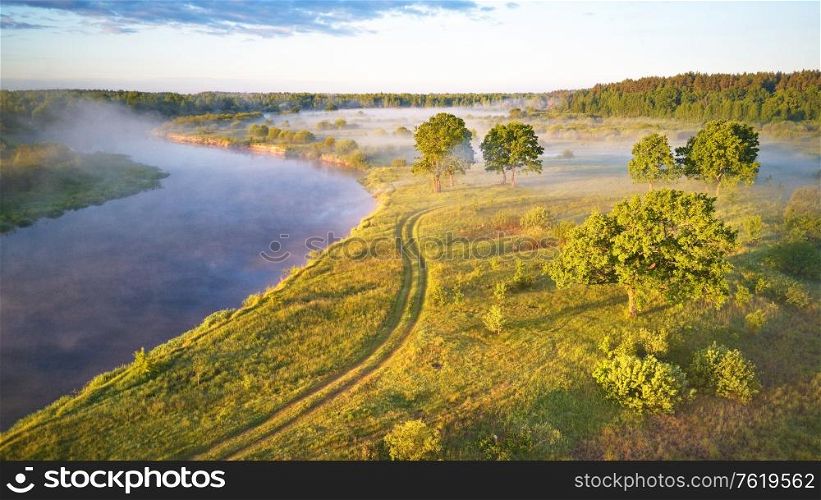 Summer misty sunrise on meadow. Country road on green fields riverbank. Sunlight on large oak trees grove in morning. Aerial view, Belarus, Berezina river