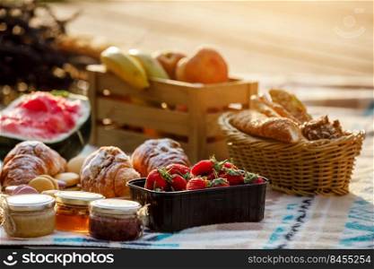 summer picnic basket with fruit and bakery on a blanket outdoors. Lunch with croissants, jam, watermelon, strawberry and fresh fruits in wooden box in the park. Copy space. summer picnic basket with fruit and bakery on a blanket outdoors. Lunch with croissants, jam, watermelon, strawberry and fresh fruits in wooden box in the park. Copy space.