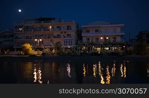 Summer resort by the sea at night with outdoor cafe, hotel and people enjoying their rest. Electric lights reflecting in water