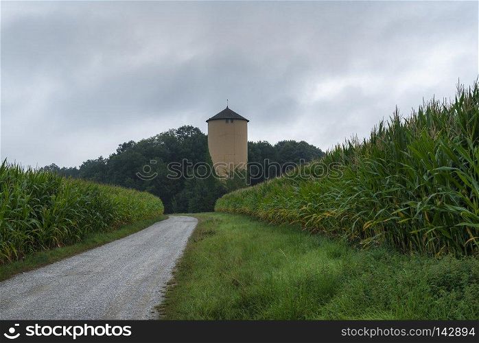 Summer rural scenery with a country road crossing green corn fields, an old tower, and the forest, on a gloomy day, near Schwabisch Hall, Germany.