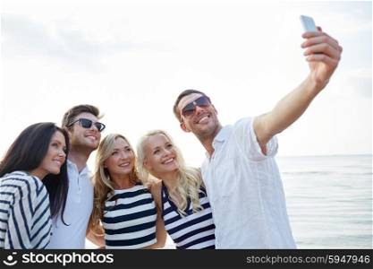 summer, sea, tourism, technology and people concept - group of smiling friends with smartphone on beach photographing and taking selfie