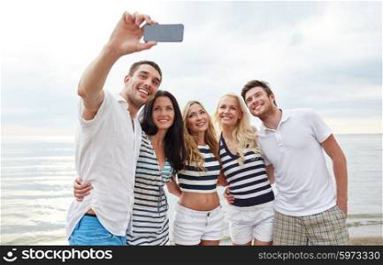summer, sea, tourism, technology and people concept - group of smiling friends with smartphone on beach photographing and taking selfie