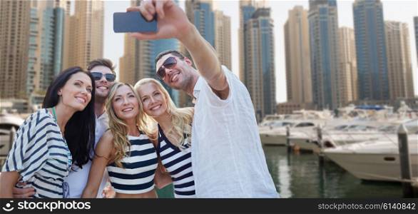 summer, sea, tourism, technology and people concept - group of smiling friends with smartphone taking selfie over dubai city harbor or waterfront background