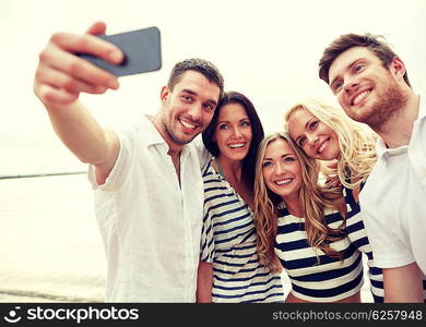 summer, sea, tourism, technology and people concept - group of smiling friends with smartphone on beach photographing and taking selfie