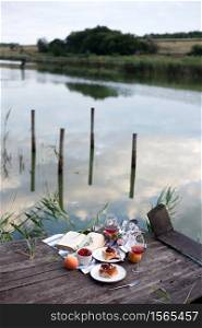 Summer still life- picnic on a wooden pier. wine, sinabones, peaches, a book with a lake view