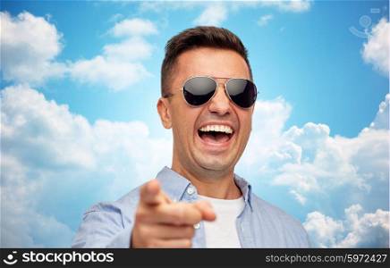 summer, style, emotions and people concept - laughing man in sunglasses pointing finger on you over blue sky and clouds background