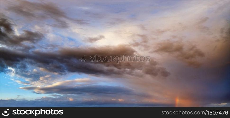Summer sunset after rain sky panorama with fleecy purple clouds and rainbow. Evening dusk good weather natural background.