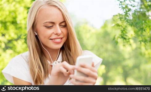 summer, technology and people concept - smiling young woman in white dress with smartphone and earphones listening to music over green natural background. woman with smartphone listening to music . woman with smartphone listening to music