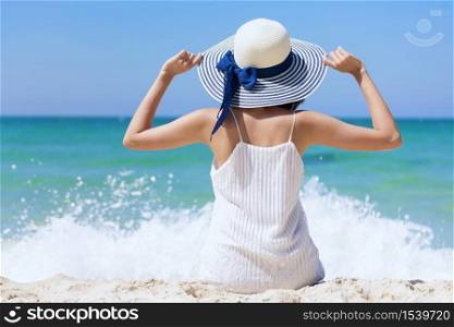 Summer time woman vacation on the beach. Cheerful woman wear summer dress and straw hats sitting on the beach look at sea. Time to relax in summer lifestyle outdoor shot on tropical island beach.