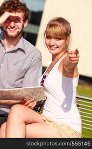 Summer tourism concept. Young tourist couple two friends on vacation in city looking up directions on map, sitting on bench at sunny day. Tourist couple in city read map