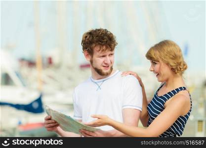 Summer travel concept. Young tourist couple on vacation standing in front of boats in marina looking up directions on map