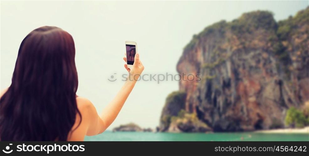 summer, travel, technology and people concept - close up of sexy young woman taking selfie with smartphone over rock on bali beach background