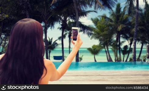 summer, travel, technology and people concept - close up of sexy young woman taking selfie with smartphone over tropical beach with palms and swimming pool background