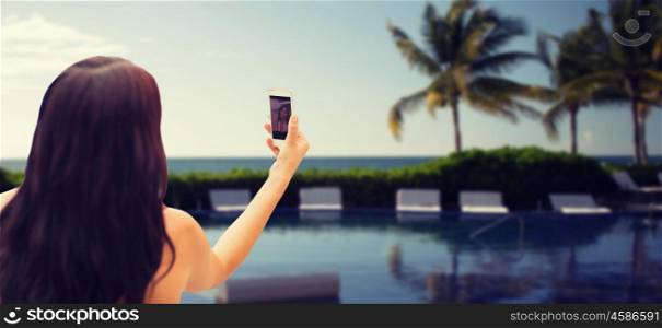 summer, travel, technology and people concept - close up of sexy young woman taking selfie with smartphone over resort beach with palms and swimming pool background