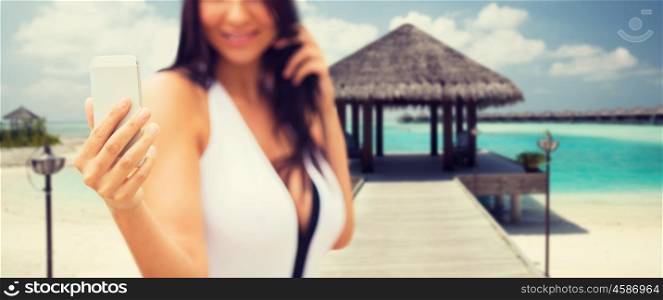 summer, travel, technology and people concept - close up of sexy young woman taking selfie with smartphone over bungalow on beach background
