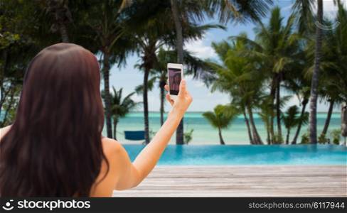 summer, travel, technology and people concept - close up of sexy young woman taking selfie with smartphone over tropical beach with palms and swimming pool background