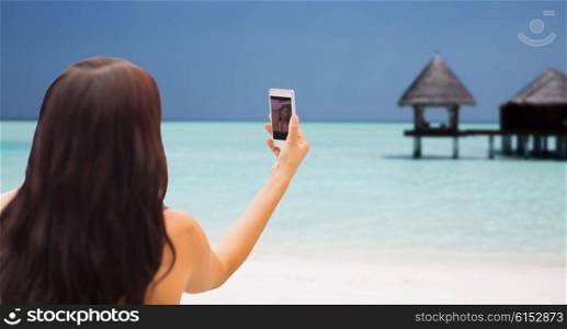 summer, travel, technology and people concept - close up of sexy young woman taking selfie with smartphone over bungalow on beach background