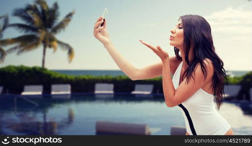 summer, travel, technology and people concept - sexy young woman taking selfie with smartphone and sending blow kiss over resort beach with palms and swimming pool background