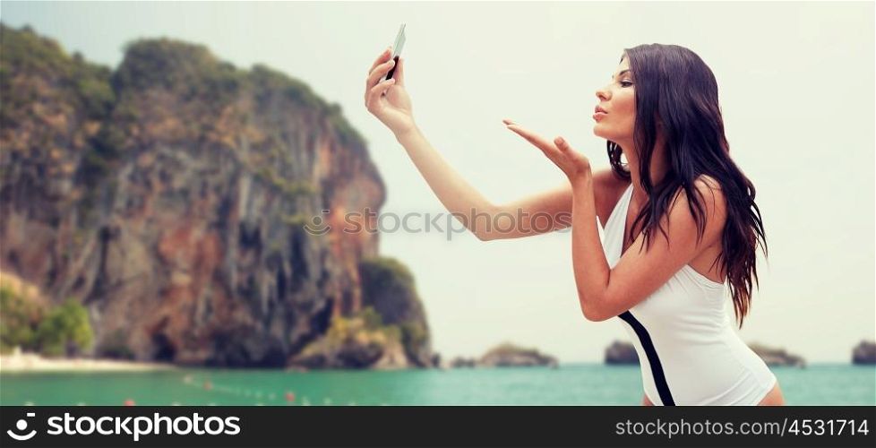 summer, travel, technology and people concept - sexy young woman taking selfie with smartphone and sending blow kiss over rock on bali beach background