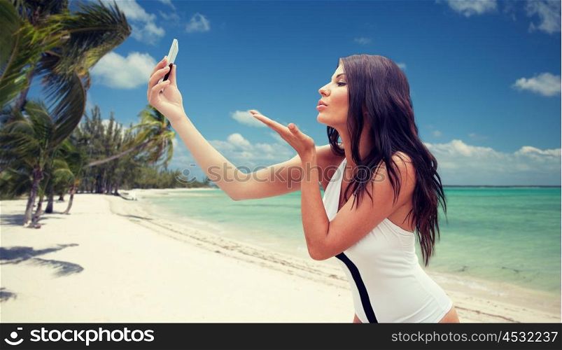 summer, travel, technology and people concept - sexy young woman taking selfie with smartphone and sending blow kiss over tropical beach with palms background