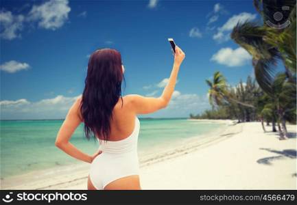 summer, travel, technology and people concept - sexy young woman taking selfie with smartphone over tropical beach with palms background