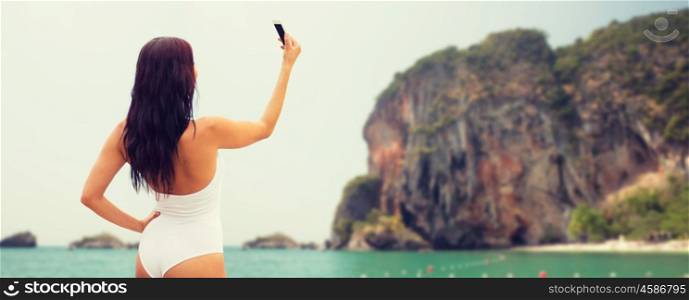 summer, travel, technology and people concept - sexy young woman taking selfie with smartphone over bali beach and rock background