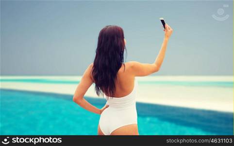 summer, travel, technology and people concept - sexy young woman taking selfie with smartphone over beach and swimming pool background