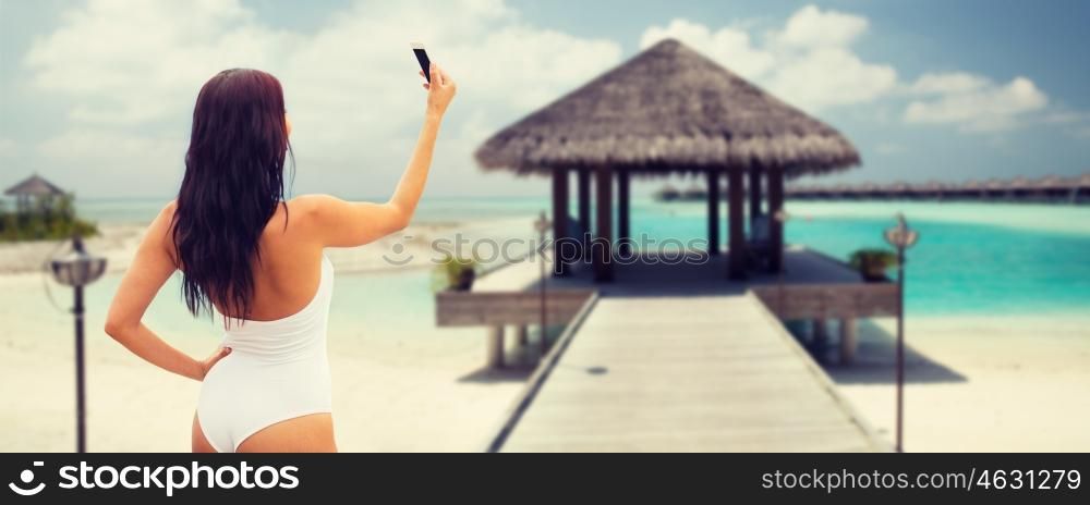 summer, travel, technology and people concept - sexy young woman taking selfie with smartphone over bungalow on beach background
