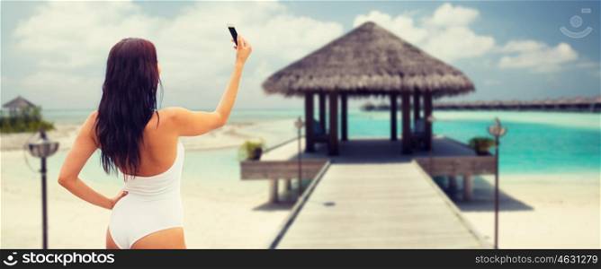 summer, travel, technology and people concept - sexy young woman taking selfie with smartphone over bungalow on beach background