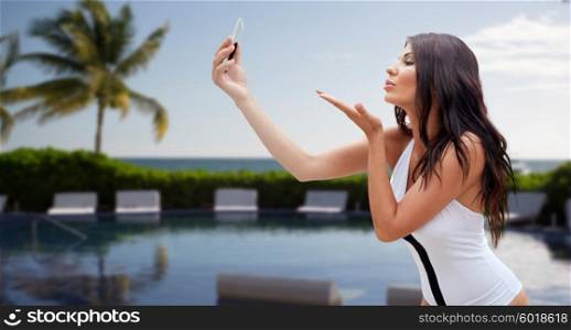 summer, travel, technology and people concept - sexy young woman taking selfie with smartphone and sending blow kiss over resort beach with palms and swimming pool background
