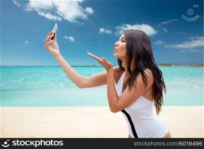 summer, travel, technology and people concept - sexy young woman taking selfie with smartphone and sending blow kiss over tropical beach over tropical beach background