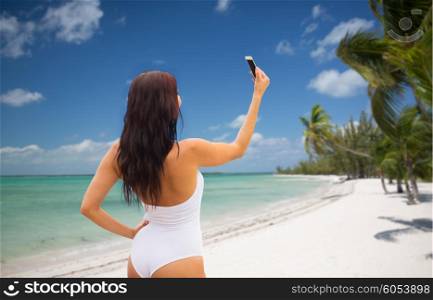 summer, travel, technology and people concept - sexy young woman taking selfie with smartphone over tropical beach with palms background