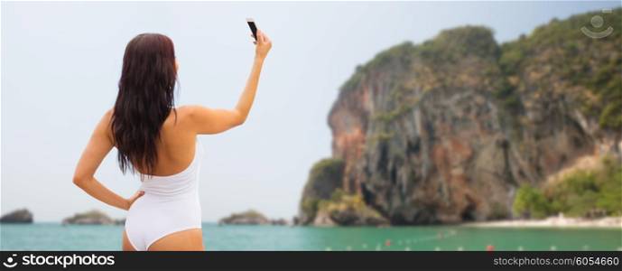 summer, travel, technology and people concept - sexy young woman taking selfie with smartphone over bali beach and rock background