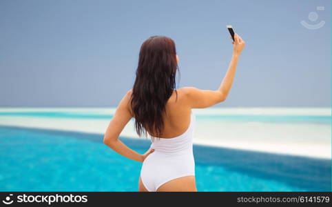 summer, travel, technology and people concept - sexy young woman taking selfie with smartphone over beach and swimming pool background