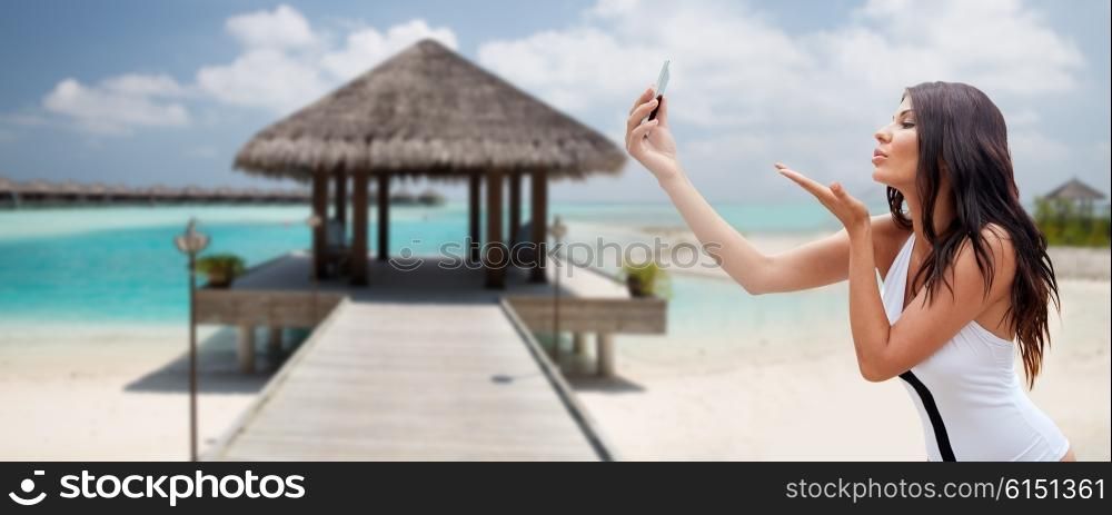 summer, travel, technology and people concept - sexy young woman taking selfie with smartphone and sending blow kiss over bungalow on beach background