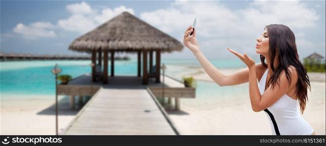 summer, travel, technology and people concept - sexy young woman taking selfie with smartphone and sending blow kiss over bungalow on beach background