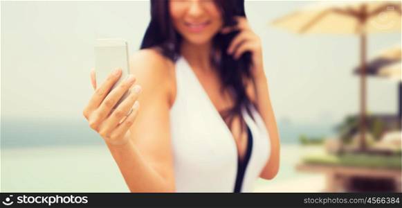summer, travel, tourism, technology and people concept - close up of sexy young woman taking selfie with smartphone over resort beach with parasols and swimming pool background