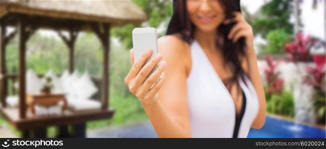 summer, travel, tourism, technology and people concept - close up of sexy young woman taking selfie with smartphone over exotic hotel resort with bungalow and swimming pool background