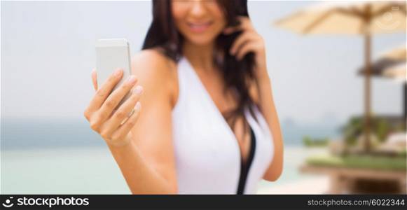 summer, travel, tourism, technology and people concept - close up of sexy young woman taking selfie with smartphone over resort beach with parasols and swimming pool background