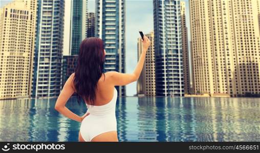 summer, travel, tourism, technology and people concept - sexy young woman taking selfie with smartphone over infinity edge swimming pool in dubai city background